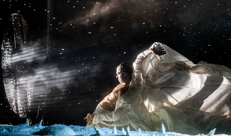 Ein Tänzer in einem fließenden Kostüm bewegt sich auf einer Bühne mit gefrorenen Elementen vor einem sternenübersäten Hintergrund.