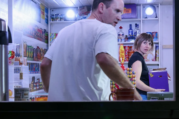 Two people in a small shop, surrounded by shelves of candy and snacks. One person holds a purple book.