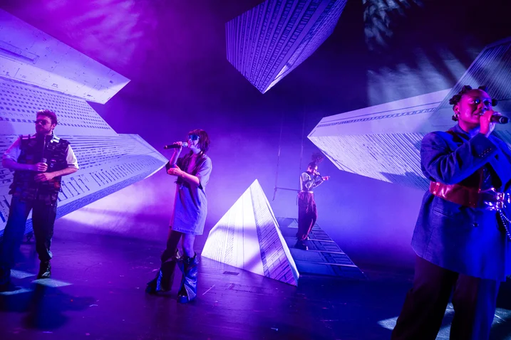 Musicians perform on a stylishly lit stage with geometric shapes in the background. Blue and purple lights dominate the scene.