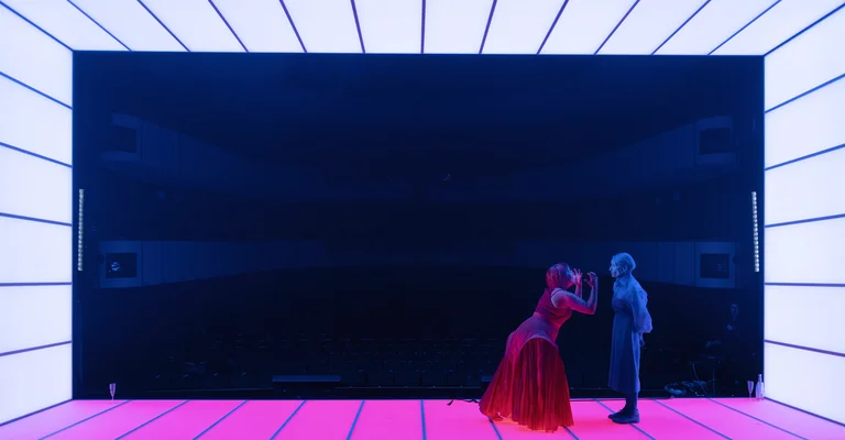 Two dancers in a futuristic, illuminated space; one in red, the other in blue, contrasting against a glowing floor.