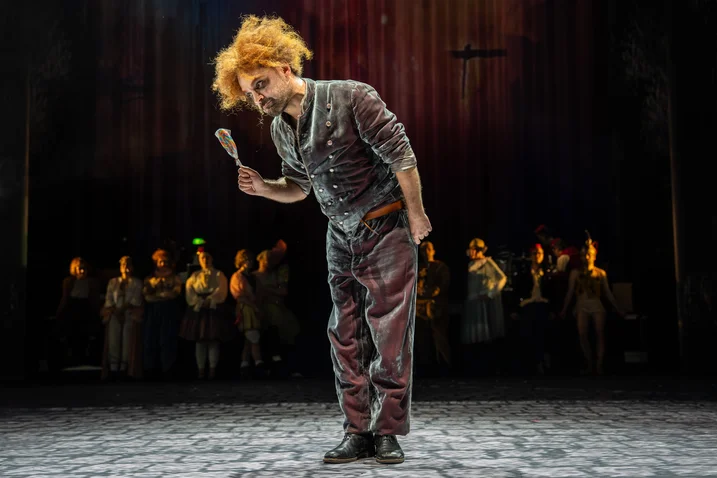 An actor with wild, red hair bows on stage. Blurred figures stand in the background under spotlight.