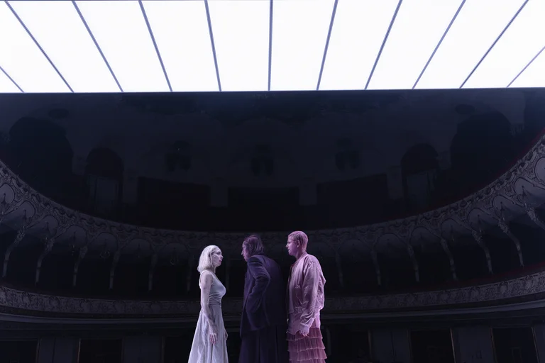 Three people stand on a theater stage. The background is dark, illuminated by a bright overhead light.