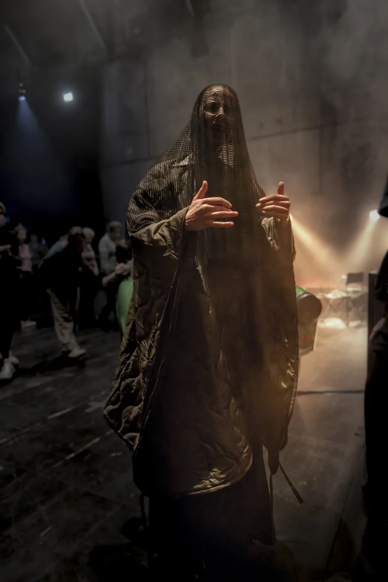 Ein Performer in einem dunklen, atmosphärischen Raum trägt ein langes Gewand und hat einen Schleier vor dem Gesicht.