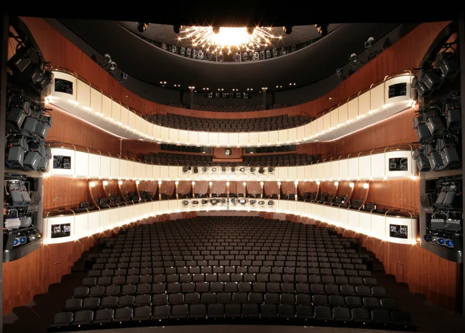 Ein modernes Theater mit mehreren Sitzreihen und Balkonen. Die elegante Innenarchitektur umfasst Holzakzente und eine große Deckenleuchte.