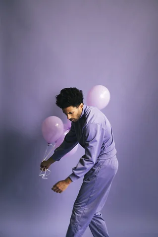 A person in a lavender outfit holds pink balloons against a lavender background. The scene feels contemplative.