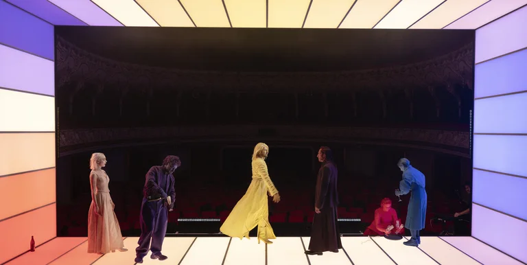 Six people stand in a room with rainbow-colored stripes on the walls. They wear costumes in various colors.