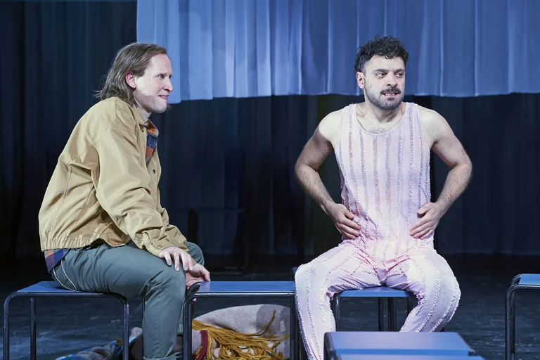 Zwei Männer sitzen auf der Bühne, einer in einem gestreiften Outfit. Sie scheinen in ein Gespräch vertieft zu sein.