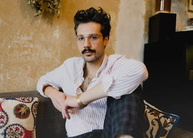 A man with glasses and a mustache sits casually on a sofa against a rustic wall.
