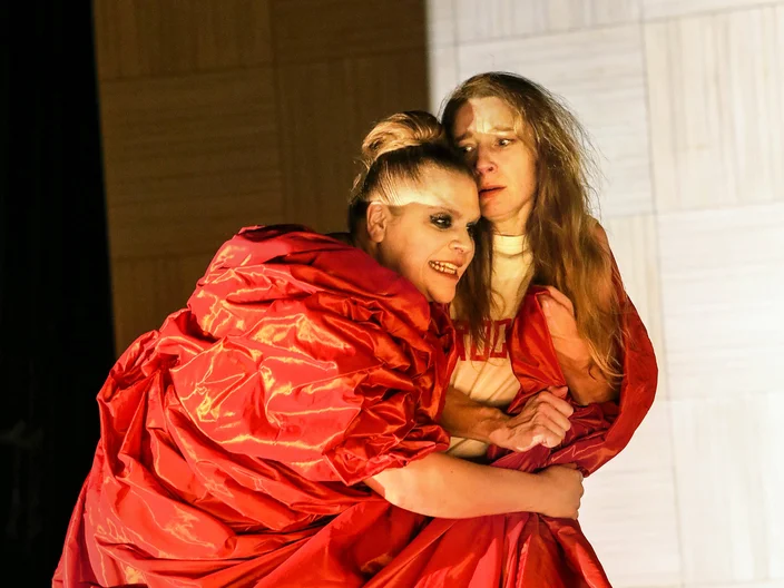 Zwei Darstellerinnen in leuchtend roten Kostümen auf einer Bühne. Sie agieren emotional, umgeben von einem minimalistischen Bühnenbild.