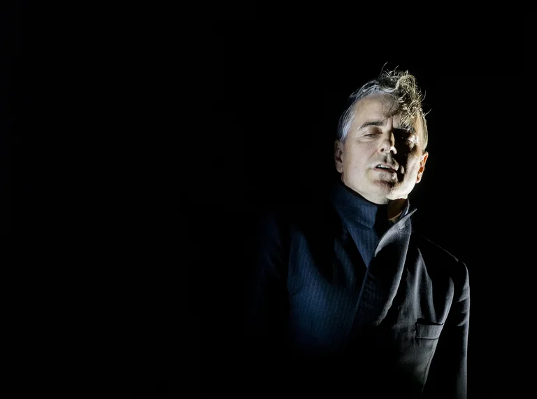 Ein Mann mit beleuchtetem Gesicht steht im Dunkeln, trägt dunkle Kleidung und schaut nach vorne. Dramatische Beleuchtung erzeugt geheimnisvolle Atmosphäre.