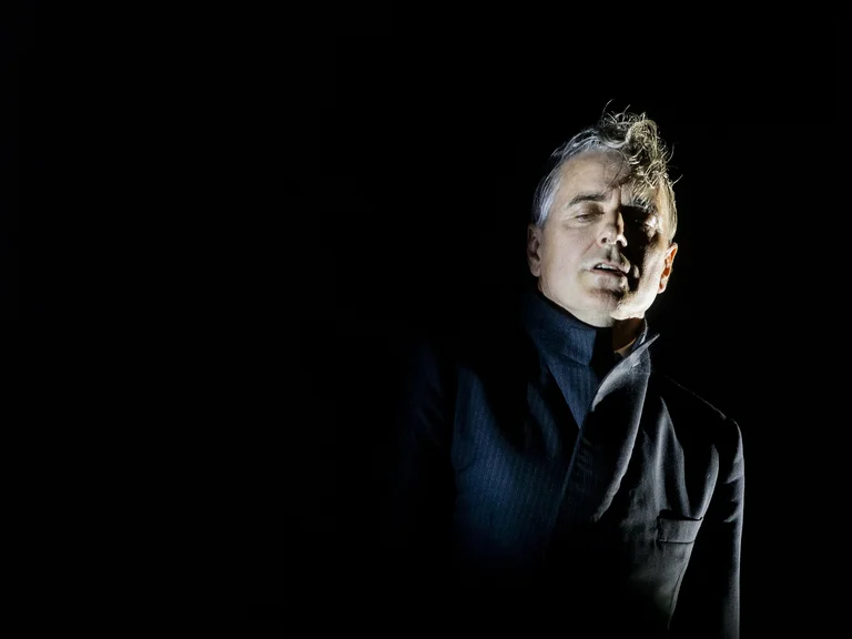 Ein Mann mit beleuchtetem Gesicht steht im Dunkeln, trägt dunkle Kleidung und schaut nach vorne. Dramatische Beleuchtung erzeugt geheimnisvolle Atmosphäre.