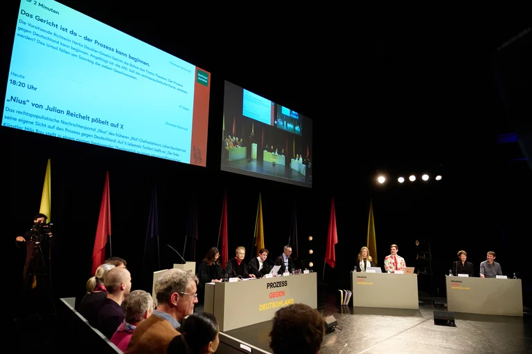 Eine Podiumsdiskussion mit mehreren Personen auf der Bühne. Im Hintergrund sind Fahnen und ein großer Bildschirm sichtbar.