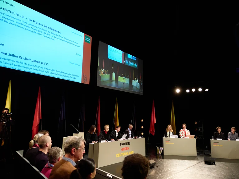 Eine Podiumsdiskussion mit mehreren Personen auf der Bühne. Im Hintergrund sind Fahnen und ein großer Bildschirm sichtbar.