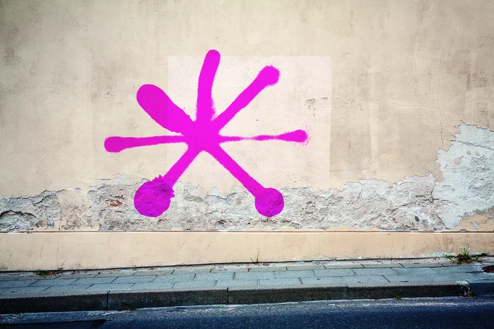 A bright pink graffiti symbol on a weathered beige wall above a sidewalk. The wall shows signs of wear.