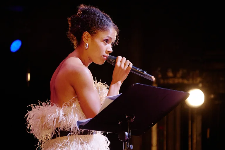 Schauspielerin Gloria Odosi in einem eleganten, federbesetzten Kleid hält ein Mikrofon und singt auf der Bühne. Hintergrund im Scheinwerferlicht.