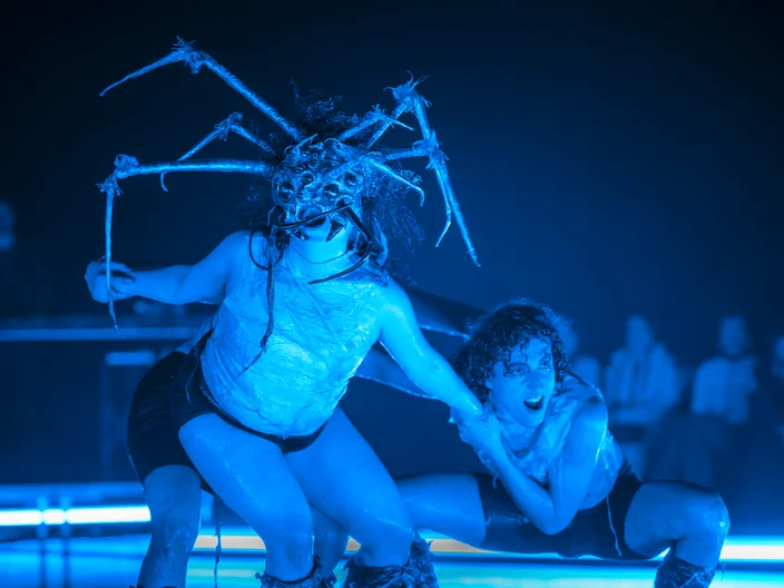 Zwei Tänzer performen auf der Bühne in blauem Licht. Einer trägt einen Kopfschmuck mit spinnartigen Strukturen. Publikum im Hintergrund.
