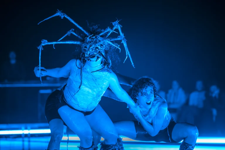 Zwei Tänzer performen auf der Bühne in blauem Licht. Einer trägt einen Kopfschmuck mit spinnartigen Strukturen. Publikum im Hintergrund.