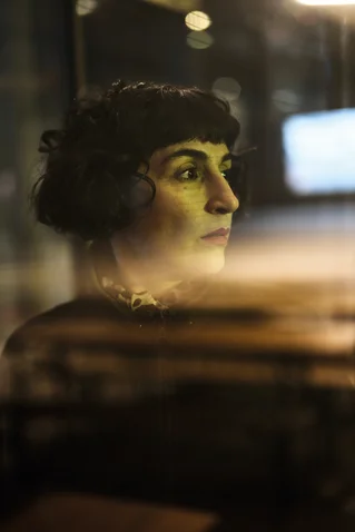 Eine Frau mit dunklen Locken, reflektiert in einem Fenster. Sie schaut ernst in die Ferne bei schwachem Licht.