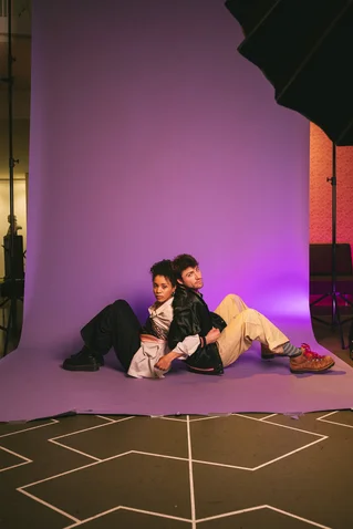 Two people sit back-to-back on a purple backdrop. They are dressed in casual, modern attire and appear relaxed.