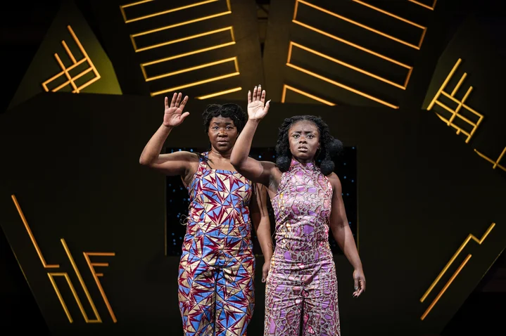 Two women in colorful patterned dresses raise their hands on stage. Geometric light patterns are visible in the background.