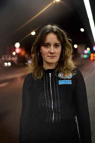 Eine Frau steht bei Nacht auf einer Straße, beleuchtet von Straßenlaternen und Lichter der vorbeifahrenden Autos.