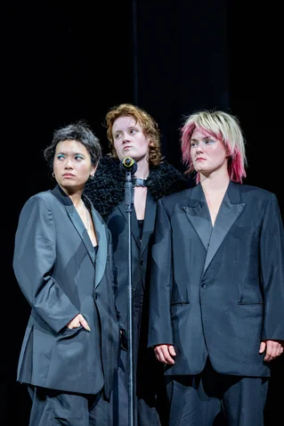 Three people in oversized suits stand on stage. They appear determined and have distinct hairstyles.