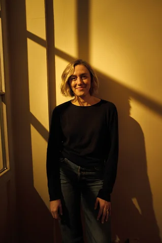 Frau steht in einem Raum mit Schattenmuster an der Wand. Sie trägt Jeans und ein dunkles Oberteil.