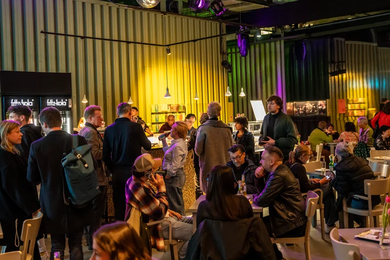 Crowd of people in a well-lit café or restaurant, standing and sitting at tables, engaging in lively conversation.