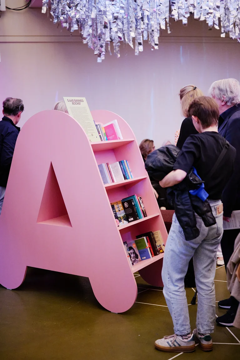 Menschen betrachten Bücher auf einem rosa, A-förmigen Regal. Das Regal ist modern gestaltet und gut beleuchtet.