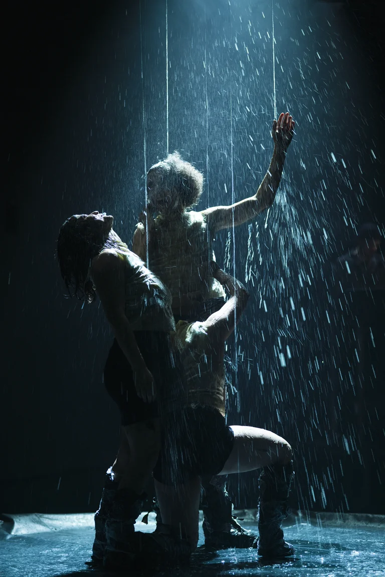 Drei Tänzer stehen unter strömendem Wasser auf einer dunklen Bühne, beleuchtet von einem dramatischen Spotlicht von oben.