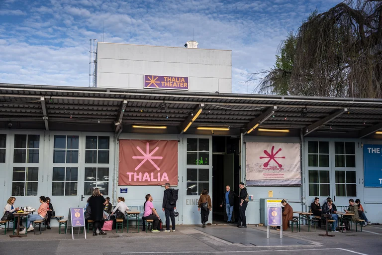 People sit and stand outside a building with Thalia banners. There's a lively, social atmosphere.