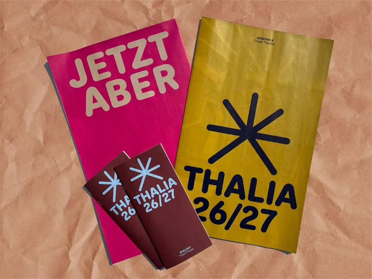 Ein rosa Plakat mit der Aufschrift „JETZT ABER“ und ein gelbes Plakat mit „THALIA 26/27“ auf braunem Hintergrund.
