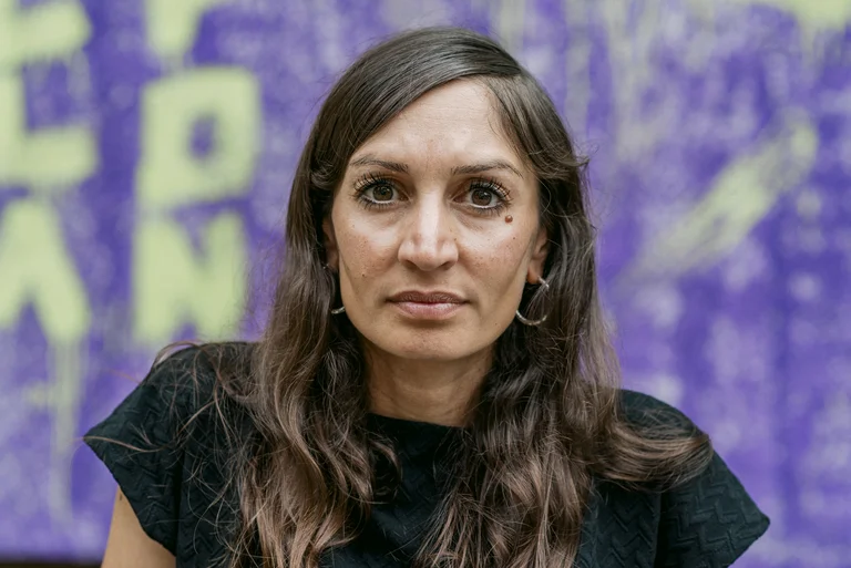 Eine Person mit langen Haaren, die ein schwarzes Oberteil trägt, steht vor einem lilafarbenen Hintergrund mit gelber Schrift.