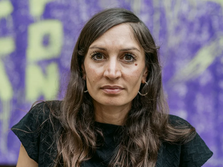 Eine Person mit langen Haaren, die ein schwarzes Oberteil trägt, steht vor einem lilafarbenen Hintergrund mit gelber Schrift.