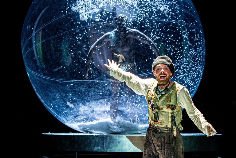 Ein Schauspieler steht vor einer großen gläsernen Kugel mit Wasser. Im Hintergrund tritt eine Person im Wasser auf.