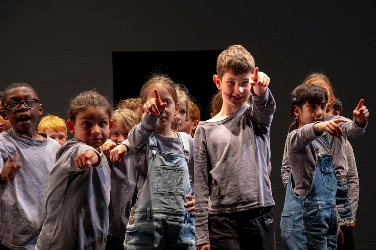 Eine Gruppe von Kindern zeigt mit ausgestrecktem Zeigefinger auf die Kamera. Sie tragen legere Kleidung und stehen vor dunklem Hintergrund.