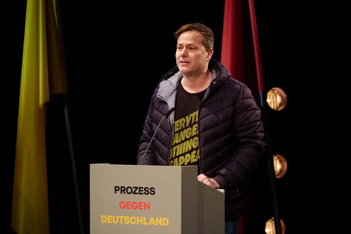 Ein Mann steht an einem Rednerpult mit der Aufschrift "Prozess gegen Deutschland". Im Hintergrund sind bunte Vorhänge zu sehen.
