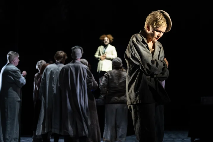 A theatrical scene with several actors in dark clothing. A figure with clown-like hair stands in the background.