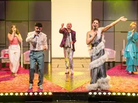 Five performers on a colorful stage passionately sing into microphones, wearing elaborate, colorful costumes under bright lighting.