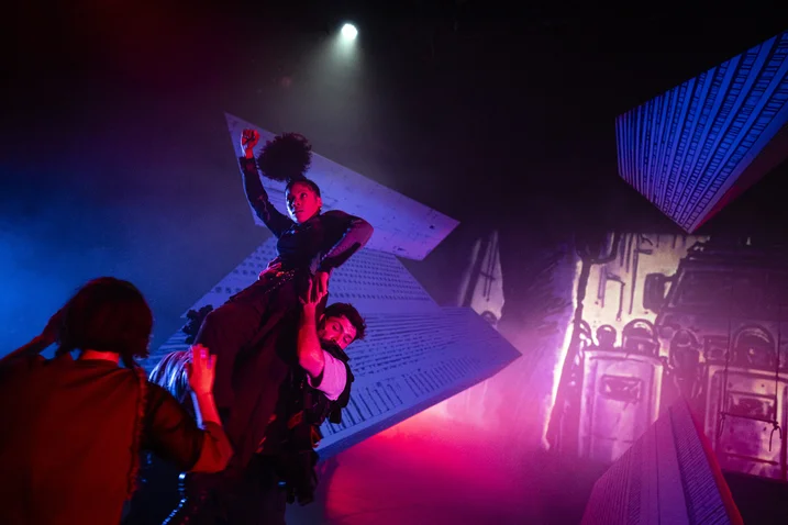 Dynamic dance scene in a colorful, futuristic setting with red and blue lighting. Dancers in expressive poses.