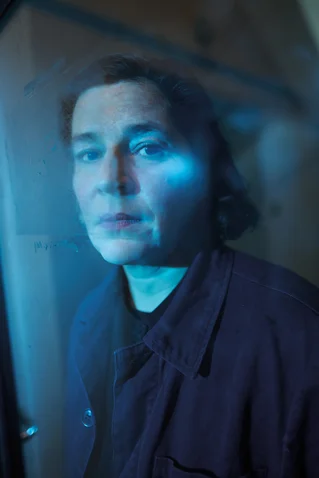 Ein Mensch steht hinter einer Glasscheibe, das Licht wirft blaue Reflexionen auf das Gesicht und die Umgebung.