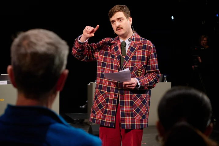 Eine Person in einem karierten Mantel hält eine Rede auf einer Bühne. Zuschauer sehen aufmerksam zu.