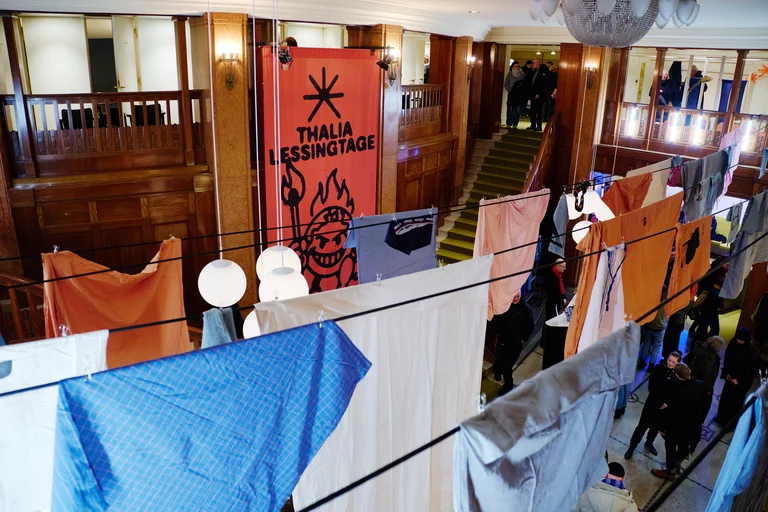 Eine Kunstausstellung mit Stoffbahnen und einem großen Banner, das die Worte "Theater Lessingtage" zeigt. Menschen schlendern durch die Ausstellung.