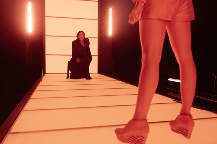 Two people in a futuristically lit corridor. One sits dressed in black, the other stands in stylish clothing and high heels.
