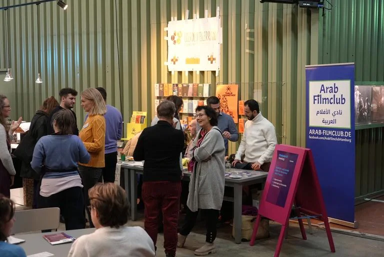 Menschen versammeln sich um einen Büchertisch auf einer Literaturveranstaltung. Ein Schild mit "Arab Literature" ist sichtbar.