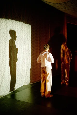 Two performers stand in front of a wall, lit to cast visible shadows. The scene creates a theatrical atmosphere.