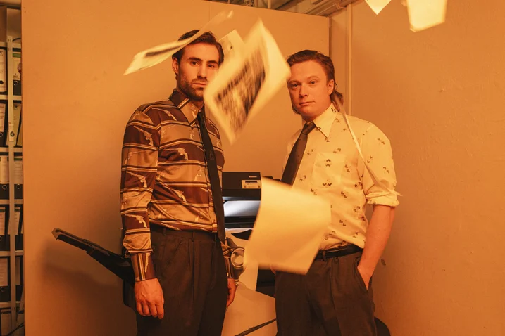 Two men in ties stand by a copier. Papers are flying, creating a chaotic office atmosphere.