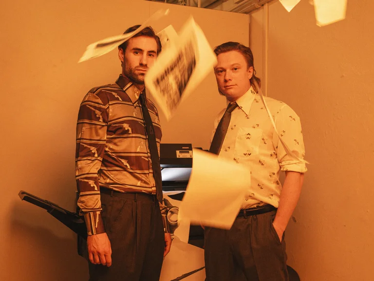 Two men in ties stand by a copier. Papers are flying, creating a chaotic office atmosphere.