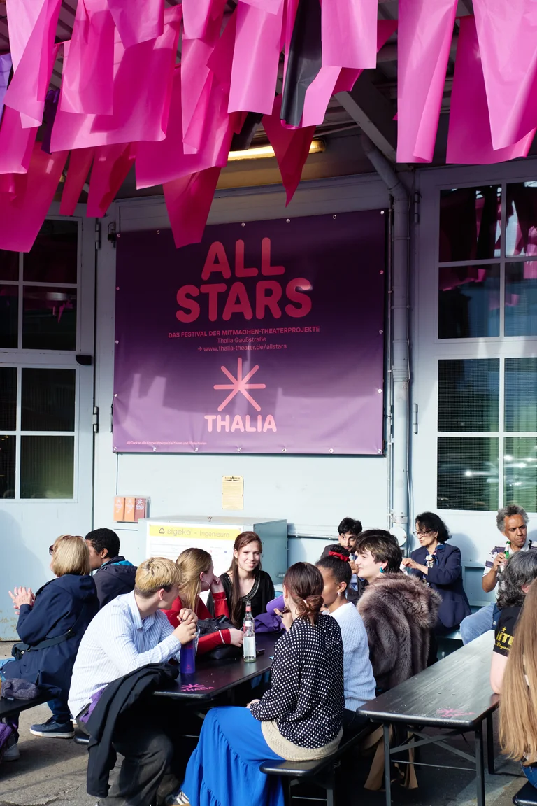 Menschen sitzen an Tischen unter rosa Dekoration. Im Hintergrund ein Schild mit "ALL STARS" und "THALIA"-Logo.