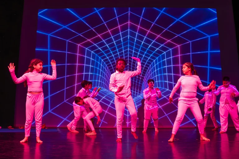 Eine Gruppe von Tänzern in futuristischen Kostümen tanzt vor einem leuchtenden, geometrischen Hintergrund in blau und pink.